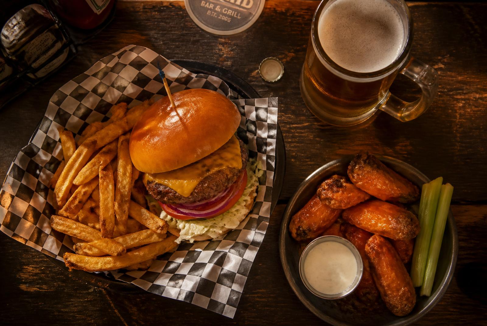 Cheeseburger with fries in a basket beside hot wings and a cold beer